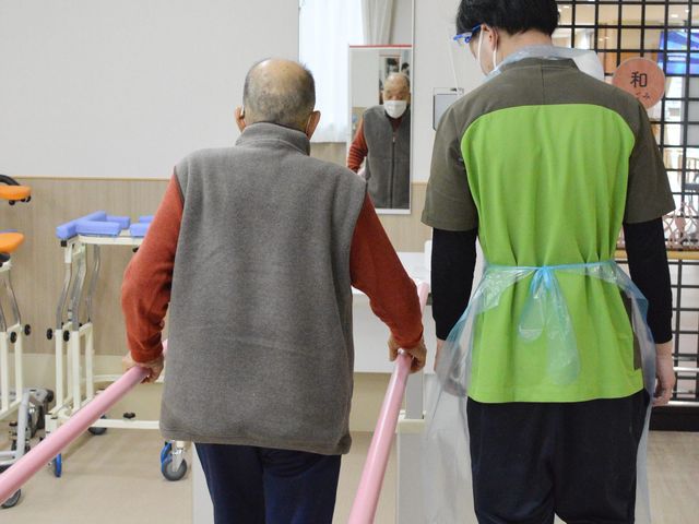 歩行補助の様子 機能訓練室 自然光が差し込む場所で、スタッフが入居者の歩行訓練を丁寧にサポートしています。