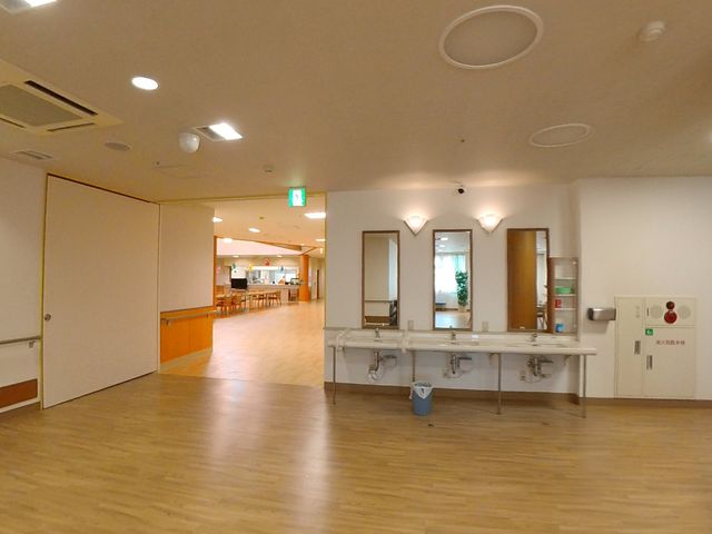 広々とした廊下の様子 食堂前スペース 床の明るい木目調と柔らかな照明が温かな印象を与える廊下です。通路の広さが余裕をもって移動できるように配慮されています。
