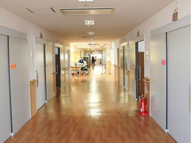 明るい廊下 通路 広々とした通路は日差しが心地よく、安心して歩ける空間を提供しています。遠くには車椅子を使用されている方が見え、安全に配慮された設計です。