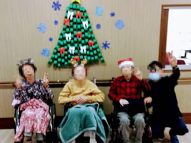 クリスマス気分の居室 クリスマスイベント 壁に飾られたクリスマスツリーの前で、利用者が笑顔でポーズを取っています。セーターや帽子も季節感を感じさせ、温かな雰囲気が伝わってきます。