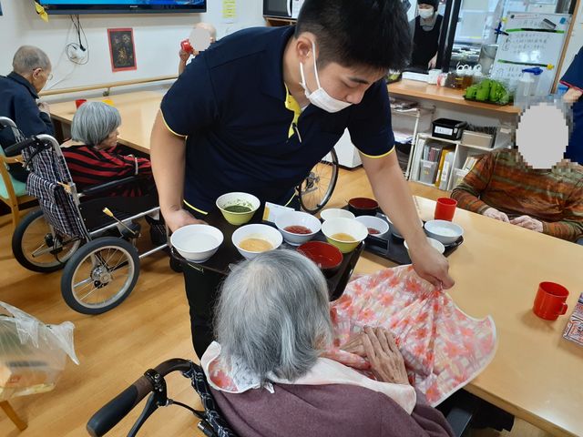 居室での昼食時 ダイニングルーム スタッフが手厚くサポートしながら、落ち着いた雰囲気の中で昼食をとる様子が伝わります。車椅子の方もゆったりと対応できる配慮が見受けられます。