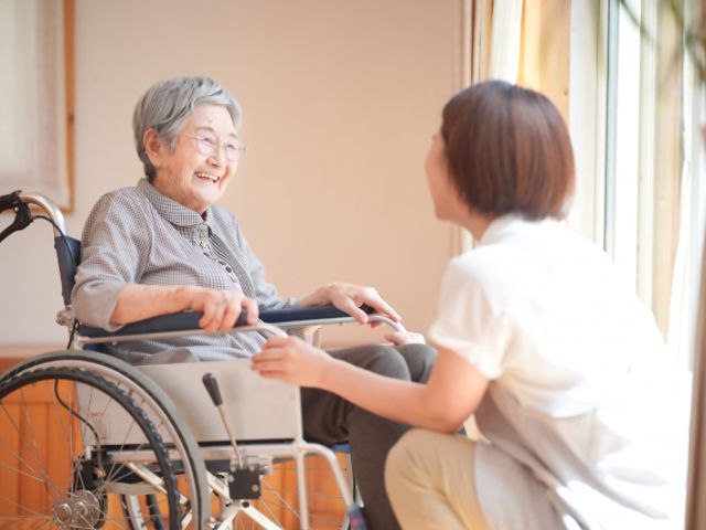 車椅子の利用者と介護士の交流 居室 明るい空間で介護士が利用者と優しくコミュニケーションを取っています。自然光が差し込む居室です。