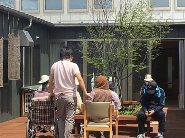 中庭でのひととき テラス席 明るい日差しが差し込む中庭で、利用者が心地よい空気を感じながらくつろぐ光景です。スタッフが穏やかに支援している様子も伺えます。