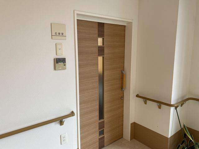 安心の個室入口 応接室 手すり沿いに設けられた、落ち着いた色合いの戸がある個室への入口です。安全に配慮された設計で、ご入居者様の快適な生活空間へと続いています。