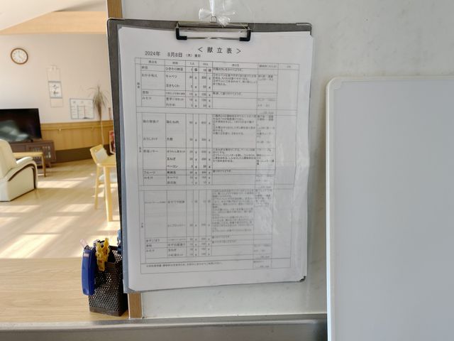 明るいエリアの掲示物 掲示物 見やすい場所に掲げられたスケジュール表です。背景にはリビングスペースが広がり、くつろぐ方々の姿が見えます。