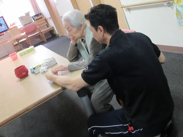 利用者と職員の交流の様子 ラウンジ 利用者が職員と向かい合ってテーブルで何かをしている様子です。安心して日々を過ごすための暖かい交流が見られます。