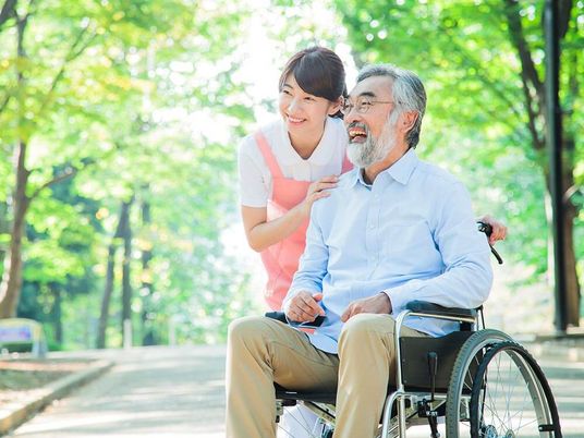 木陰を散歩する車椅子に乗った高齢男性と介助をする女性ヘルパー