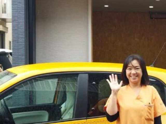 元気な挨拶の女性と車 外観 明るい表情の女性スタッフが、施設の入り口前で親しみやすい雰囲気を演出しています。