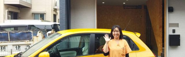 元気な挨拶の女性と車 外観 明るい表情の女性スタッフが、施設の入り口前で親しみやすい雰囲気を演出しています。