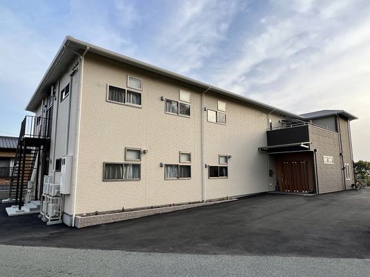 ベージュの外壁の2階建ての建物の側面には屋外階段があり、玄関には屋根が付いている。