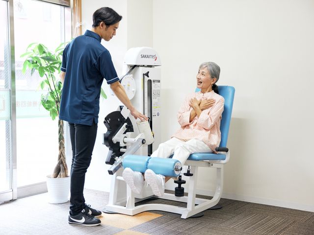 運動器具と利用者 機能訓練室 笑顔でリハビリを行う利用者様と、手厚い支援を提供するスタッフの姿が見受けられます。安心のサポート体制が整っています。