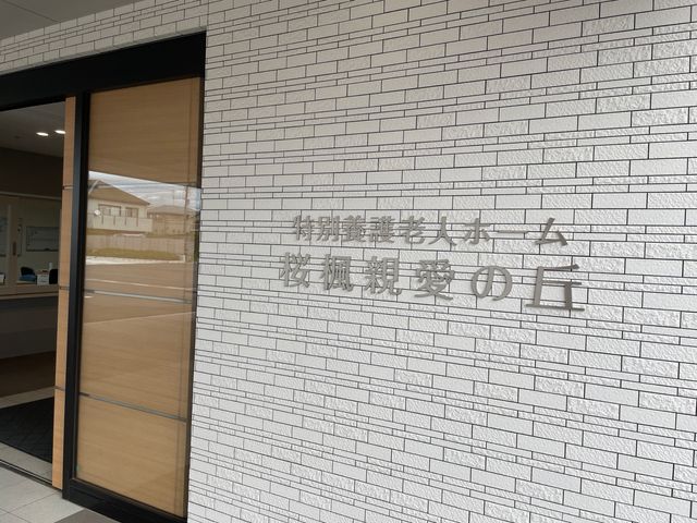 施設名のあるエントランス エントランス 白いタイル調の壁に施設名が掲げられた、落ち着いた雰囲気の入口です。大きな開口部から明るさが入り、訪れる方を迎えやすい印象です。