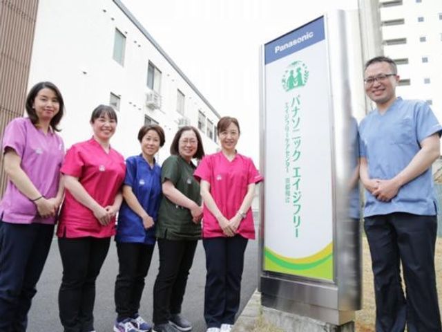 スタッフが看板の前に並ぶ外観 看板   明るい色のユニフォームを着たスタッフの皆さんが、看板のそばに並んでいます。働く人の雰囲気が伝わり、職場選びの参考にしやすいです。
