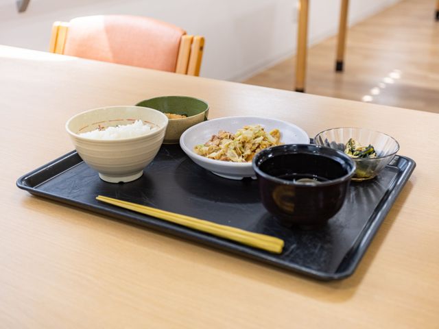 食事トレーが置かれた食堂 食堂   木目調のテーブルに、主食とおかずが並んだ食事トレーが置かれています。落ち着いた雰囲気で、食事時間の様子が伝わります。