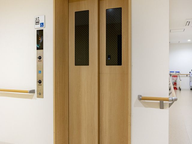 エレベーター前 エレベーター   木目調の扉が落ち着いた印象のエレベーターです。手すりが設置され、車椅子の方も移動しやすいつくりに見えます。