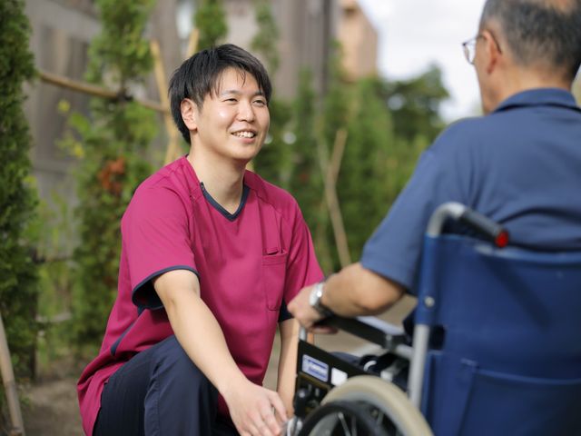 職員と利用者の会話 中庭   緑に囲まれた屋外で、職員が車椅子の方とやさしく向き合っています。落ち着いた雰囲気の中で、会話しながら過ごせそうです。