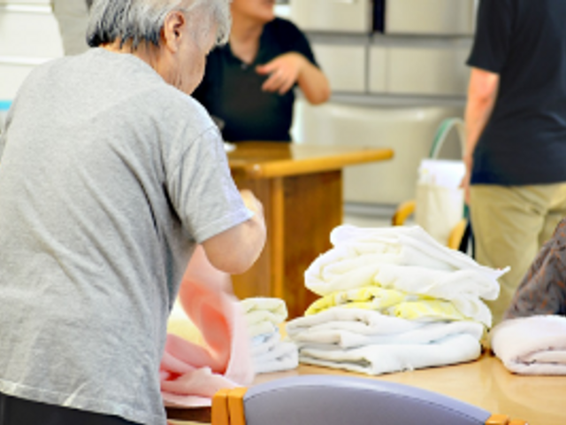 タオルをたたむ様子 多目的室 スタッフが交流しながらタオルをたたんでいます。共同作業を通じてチームワークを育みます。