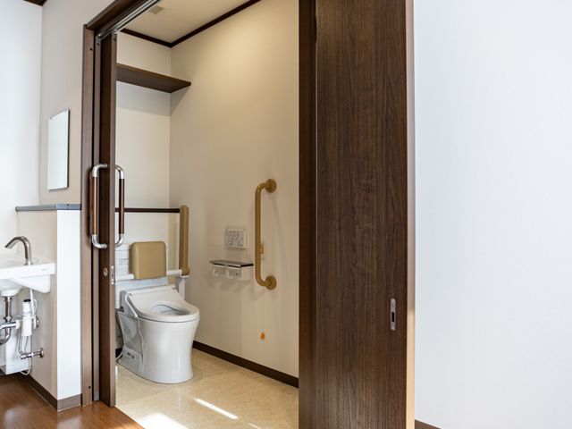 手すり付きトイレ トイレ   木目調の落ち着いた空間に、手すり付きのトイレが設置されています。ドアが大きく開くため、出入りしやすそうです。