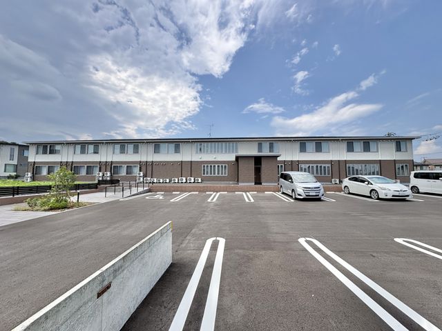 施設の外観と駐車場 外観   青空の下に、落ち着いた色合いの建物と広い駐車場が見えます。車の出入りがしやすそうで、働く方の通勤もしやすい環境に見えます。