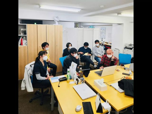 打ち合わせ中のスタッフ室 多目的室   黄色いテーブルを囲み、マスクを着けた職員が打ち合わせをしています。資料やパソコンが並び、連携の様子が伝わります。