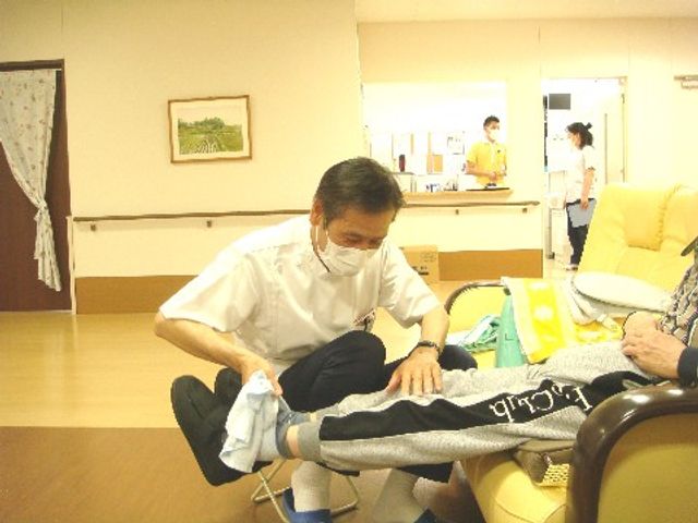 介護スタッフと利用者の様子 マッサージ スタッフが丁寧に足のケアを行っており、背景には別のスタッフが仕事をしています。安心して技術を学べる職場です。