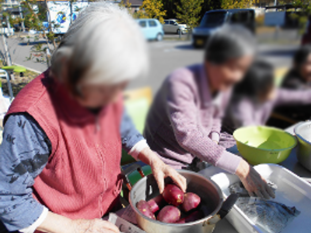 屋外での活動の光景 多目的室 元気なシニアが、屋外のテーブルで芋を使った調理を楽しんでいます。楽しそうな雰囲気が伝わります。