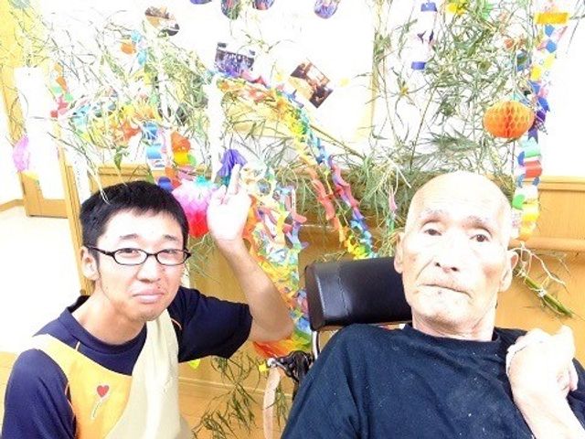 お祭り飾りと先輩と後輩 イベント 明るい装飾に囲まれた居室で、スタッフが利用者様と楽しい時を過ごしています。ハートのマークが入ったエプロンが、あたたかい雰囲気を醸し出しています。