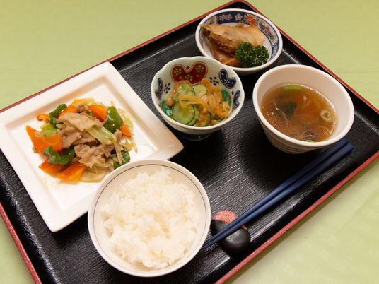 黒いトレイの上に、ご飯、スープ、彩り豊かな3種類のおかずが並べられている。豆型の箸置きに箸が載せてある。