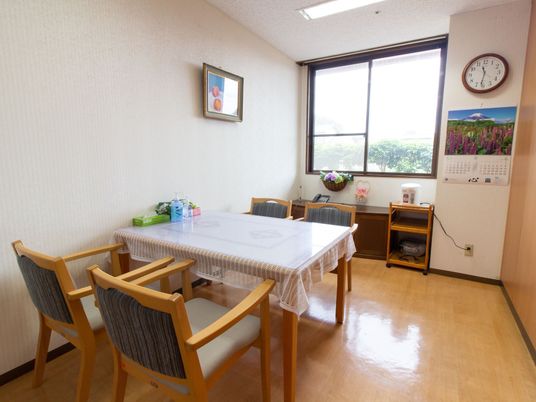 窓のある個室に、ダイニングセットが据えられている。壁面には時計やカレンダー、絵画が掛かり、窓のそばにはポットが置いてある。
