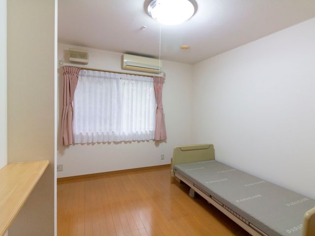 清潔な居室のインテリア 居室  広々とした居室は、明るい窓と清潔なベッドが備わり、快適な生活空間を提供しています。木目調の床が温かみを感じさせる設計です。