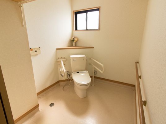淡い色あいの床のトイレ。ゆったりした広さの個室に、温水洗浄便座付きの白い便器が設置されている。各種の手すりが備えつけられている。