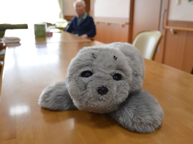 ぬいぐるみと利用者の居室 食堂 ぬいぐるみが置かれた温もりある空間で、背後にはニコリと微笑む利用者の姿があります。コミュニケーションを大切にする環境で働きませんか。