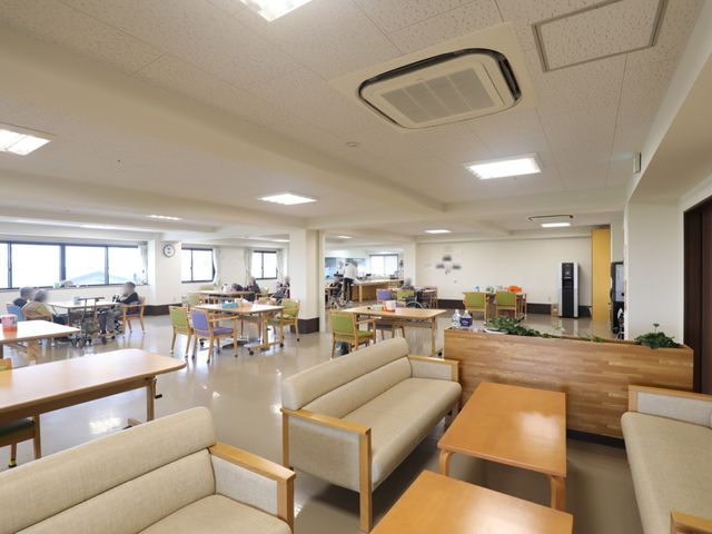 共用スペースの様子 リビング 明るく広々としたダイニングルームでは、ソファやテーブルが配され落ち着いた雰囲気でお過ごしいただけます。利用者の方々がリラックスして食事や談話を楽しんでいる様子が伺えます。
