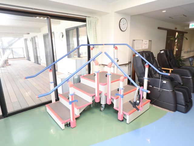 歩行訓練用階段とマッサージチェア 機能訓練室 手すり付きの階段は歩行能力の向上に役立ち、隣にはゆったりとしたマッサージチェアも完備しています。働く中で心身両面のケアを大切にする環境です。