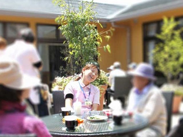 穏やかな時間の中庭 中庭 日差しの温もりを感じる中庭で、スタッフと笑顔で会話を楽しむ方々の姿が見られます。空気がきれいで心地よい屋外空間です。