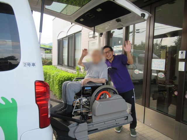 安心の送迎風景 エントランス 施設の前で、スタッフが入居者を優しく見送る場面です。温かい雰囲気が感じられます。
