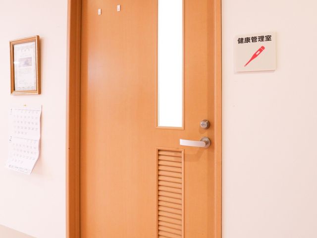 木製ドアと掲示物 健康管理室 清潔感のある廊下に設置された案内板があり、情報を手早く確認できる環境が整っています。