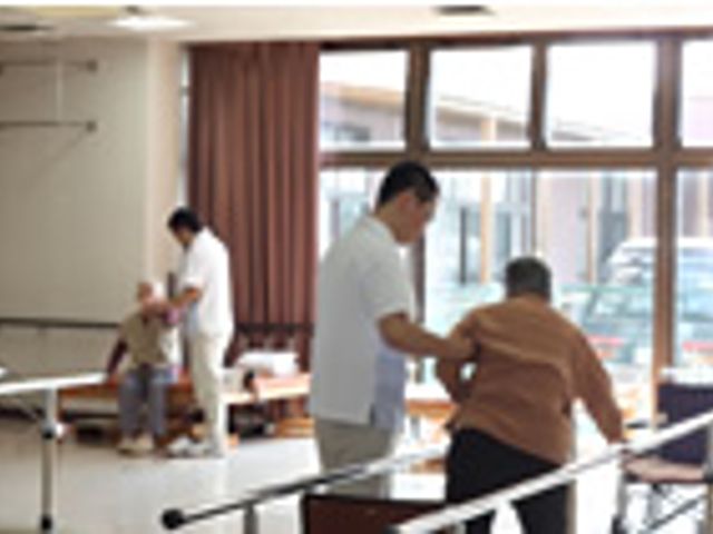 訓練支援を行う部屋 機能訓練室 自然光が入る明るい空間で、リハビリ支援が行われています。スタッフが安全にサポートしています。