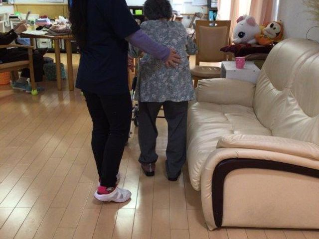 スタッフと利用者の居室 ケア 柔らかな照明の下、スタッフが優しく支えながら利用者と歩行のお手伝いをしています。明るく家庭的な雰囲気の居室で、心地よい時間を過ごしていただけます。