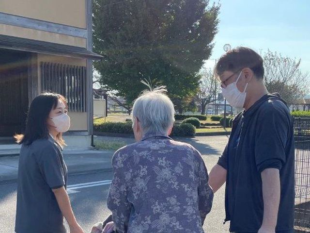 車椅子の高齢者と二人の介助者 ケア 日差しの中、スタッフが丁寧に車椅子を利用されている方のサポートをしています。心地よい環境での対話も、日々の生活に彩りを与えています。