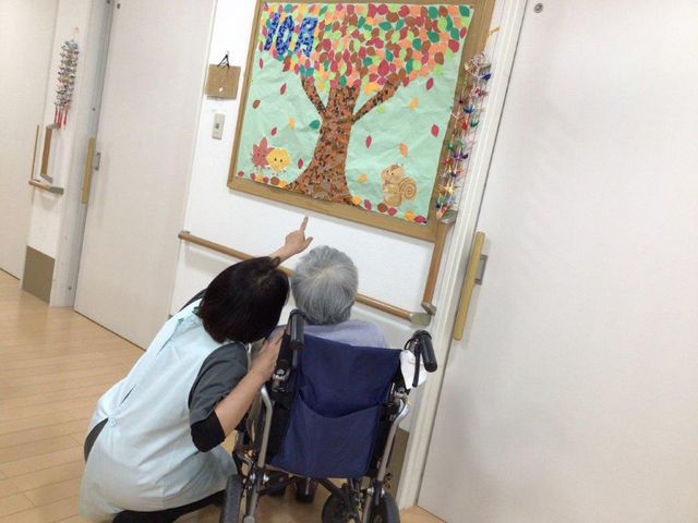 壁画を見る人とスタッフ レクリエーション 廊下にある壁画をスタッフが丁寧に説明しており、居心地の良い雰囲気を感じます。このようなふれあいが日常の一部となっています。