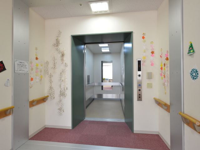 廊下にあるエレベーター エレベーター 手すりと広々とした通路が設けられた明るい廊下です。エレベーターの周りに飾られた手作りの装飾が、温かみのある雰囲気を演出しています。