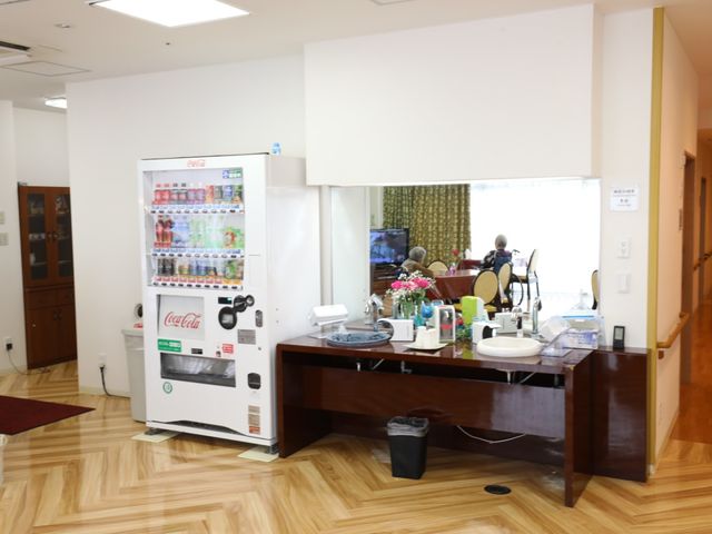 共有スペースと自動販売機 洗面スペース 清潔感のある休憩スペースで、ソフトドリンクの自動販売機が設置され、利用者の方がリラックスして過ごしています。遠くに見える窓からは自然光が差し込み、居心地の良い環境が広がっています。