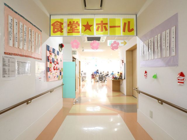 明るい廊下と食堂入口 廊下 廊下の壁には掲示物が飾られ、手前に食堂入口が見えます。遠くには明るい食堂で過ごす人々の姿が映っており、温かい雰囲気が感じられます。