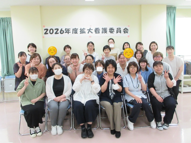 会議参加者の集合写真 多目的室 多目的室   会議に参加した職員が並んで座り、笑顔で手を振っています。広い室内で意見交換しやすい雰囲気が伝わります。