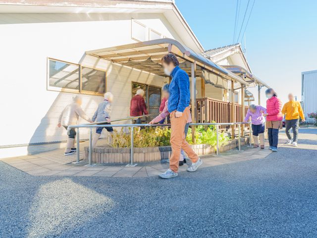 中庭で散歩する利用者たち エントランス 手すりに沿い安全に歩ける、明るい中庭。日差しを感じながら、ご高齢の方々がゆっくりと散歩をお楽しみいただけます。