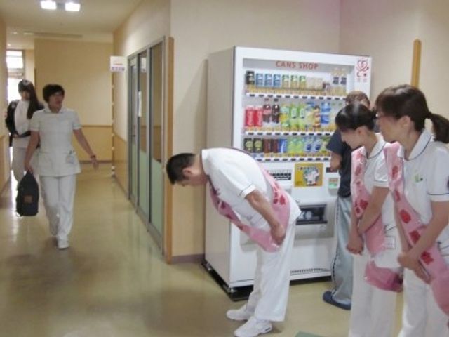 廊下と売店前の様子 廊下   明るい廊下に自動販売機があり、スタッフの方々が利用者さんに丁寧に対応しています。通路幅も確保され、動きやすさが感じられます。