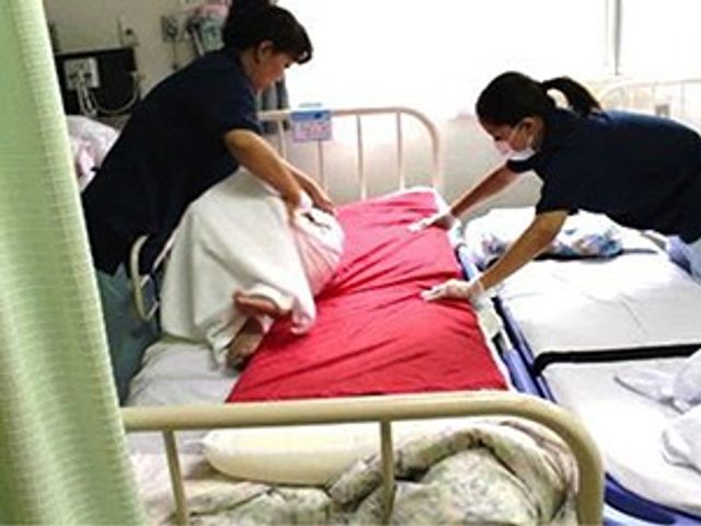 ベッド周りの介助場面 居室   ベッドで休む方を、スタッフが丁寧に整えています。日々の見守りや介助の様子が伝わる一枚です。