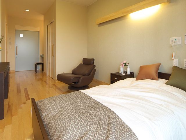 明るい居室のベッド周り 居室   木目の床が広がる居室で、ベッド横にくつろげる椅子が置かれています。落ち着いた雰囲気で、静かに過ごしやすそうです。