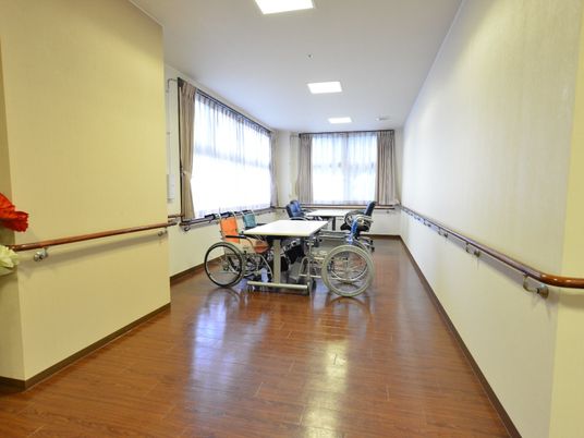 施設の写真 共有スペースは、壁2面に大きな窓があり明るい空間になっている。中央には、白い2台のテーブルと、数台の車椅子が置かれている。